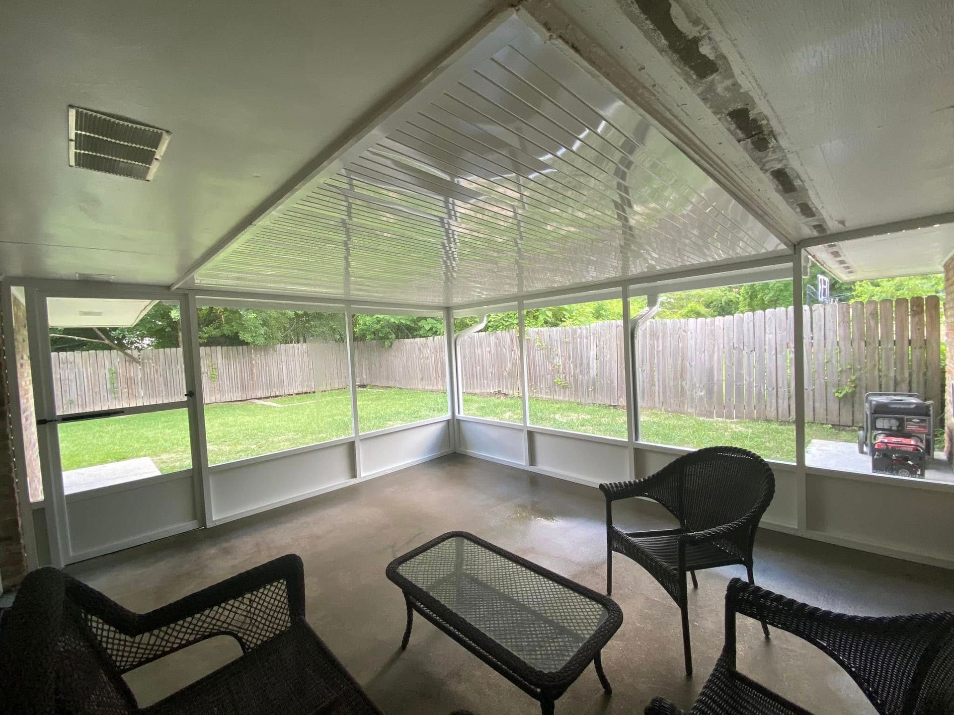 Sunroom with a clear roof and screened walls, facing a fenced backyard; furnished with wicker chairs and a glass-top table.
