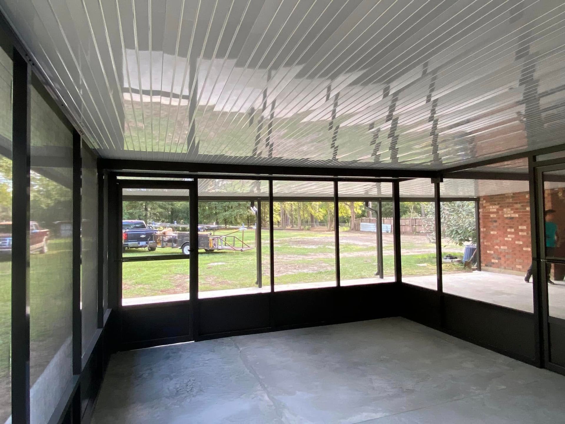 Screened-in porch with a gray floor, black frames, and a slatted ceiling. The view outside shows green grass and a truck.