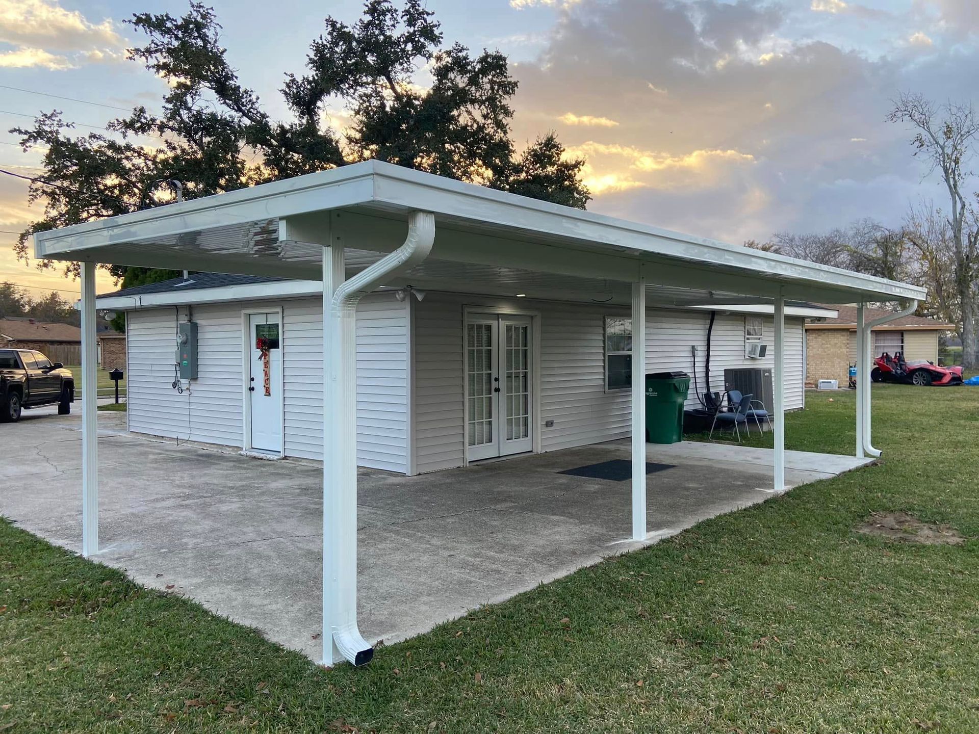 White carport over a concrete patio next to a white house; green grass and trees in the background.