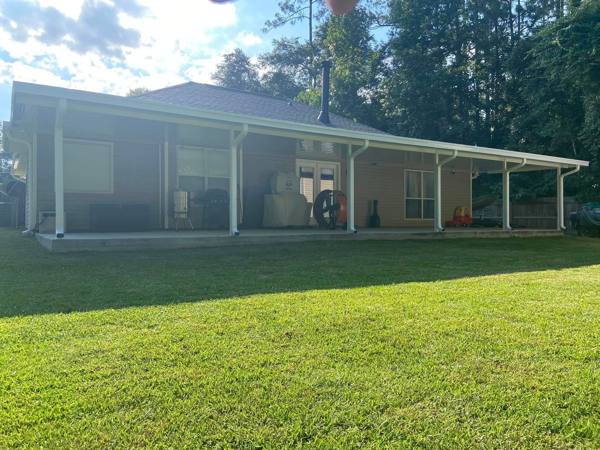 A tan house with a white-framed porch. Green grass in the foreground, trees in the background.