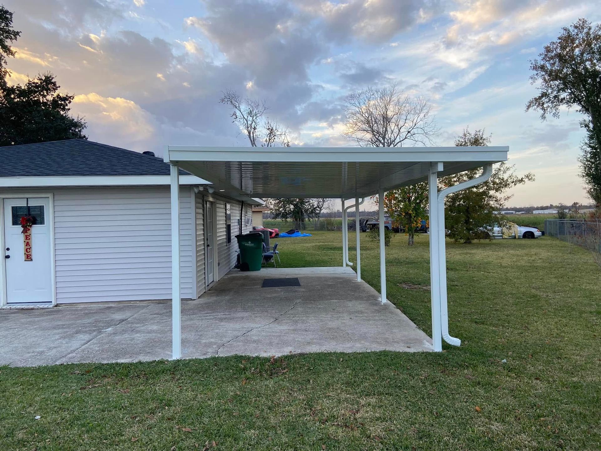 White carport attached to a small white building, with a concrete slab and green grass. Cloudy sky.
