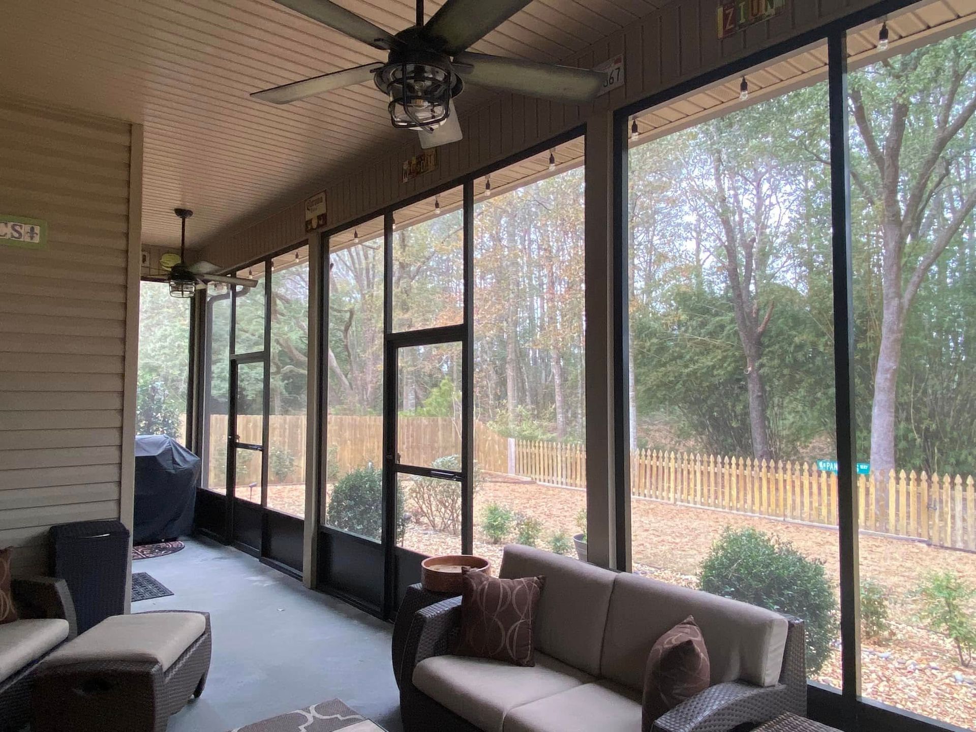 Screened porch with beige siding, ceiling fans, and a beige sofa overlooking a backyard with a wooden fence.