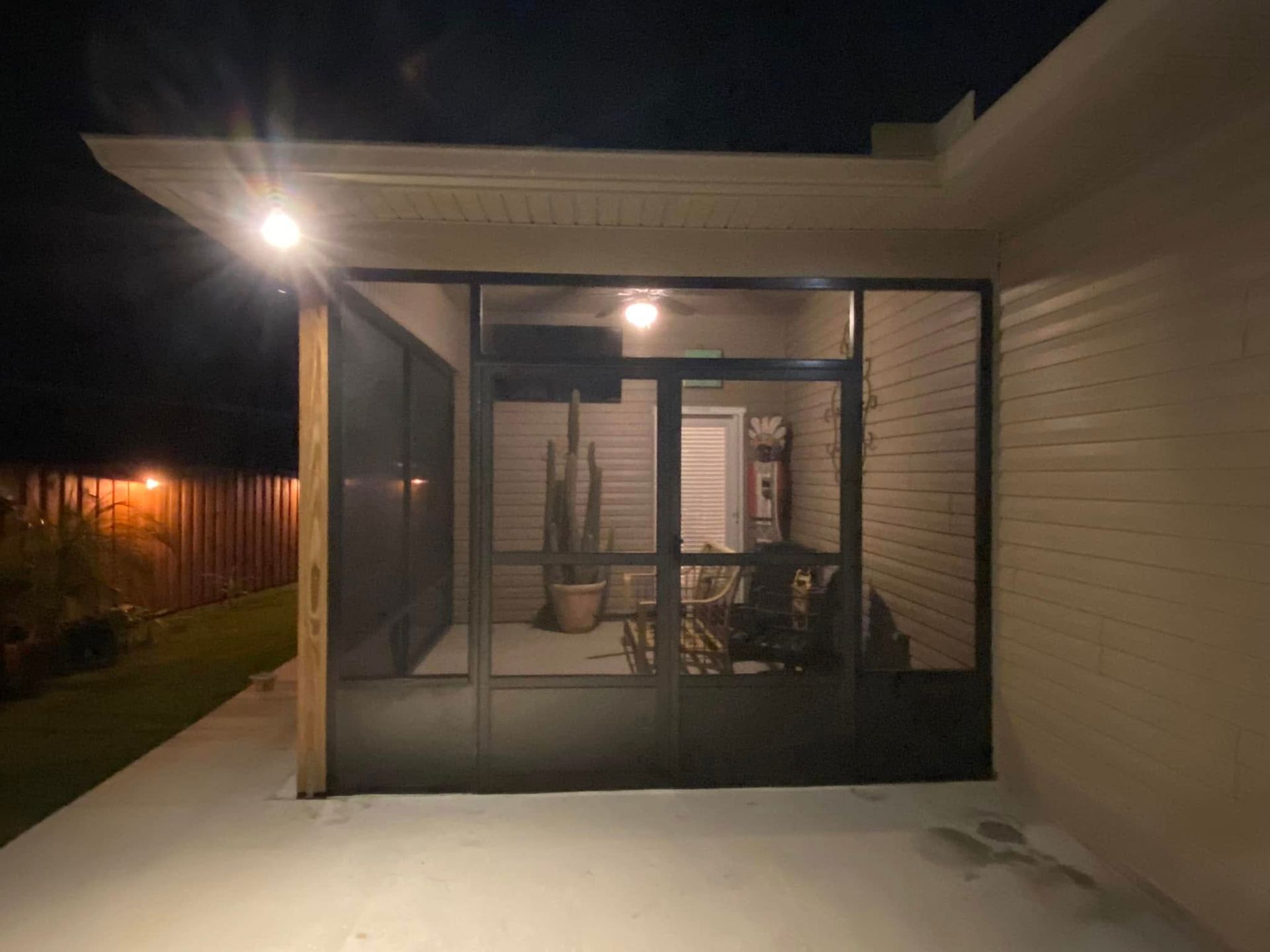 Night view of a screened porch with a light illuminating the patio. Cactus and furniture are inside.
