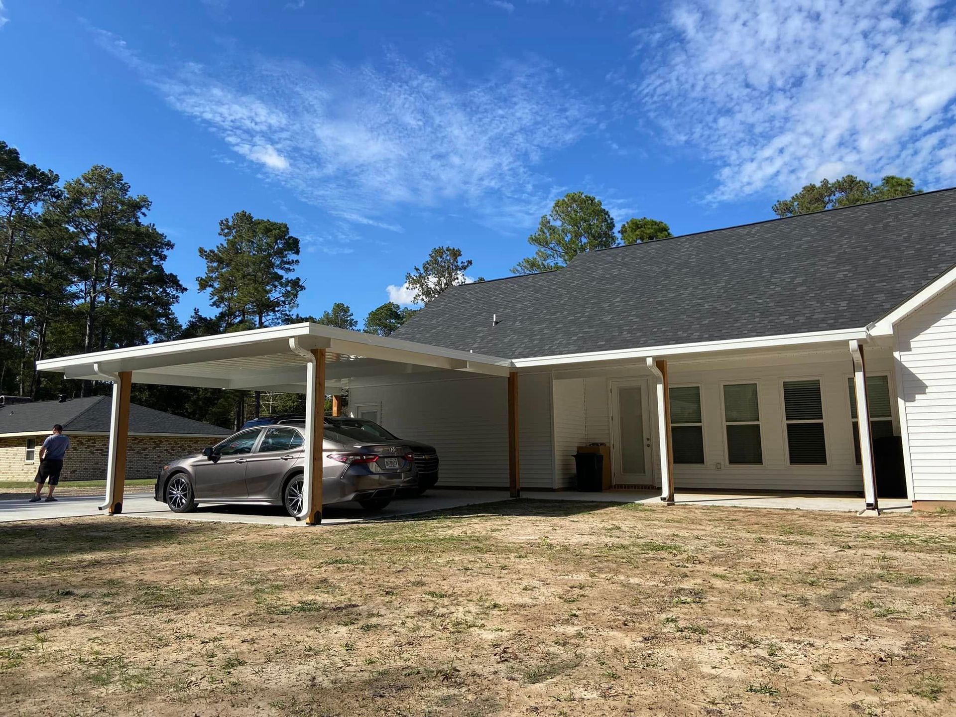 Car parked under a white carport attached to a white house. A person walks in the background. Sunny day.