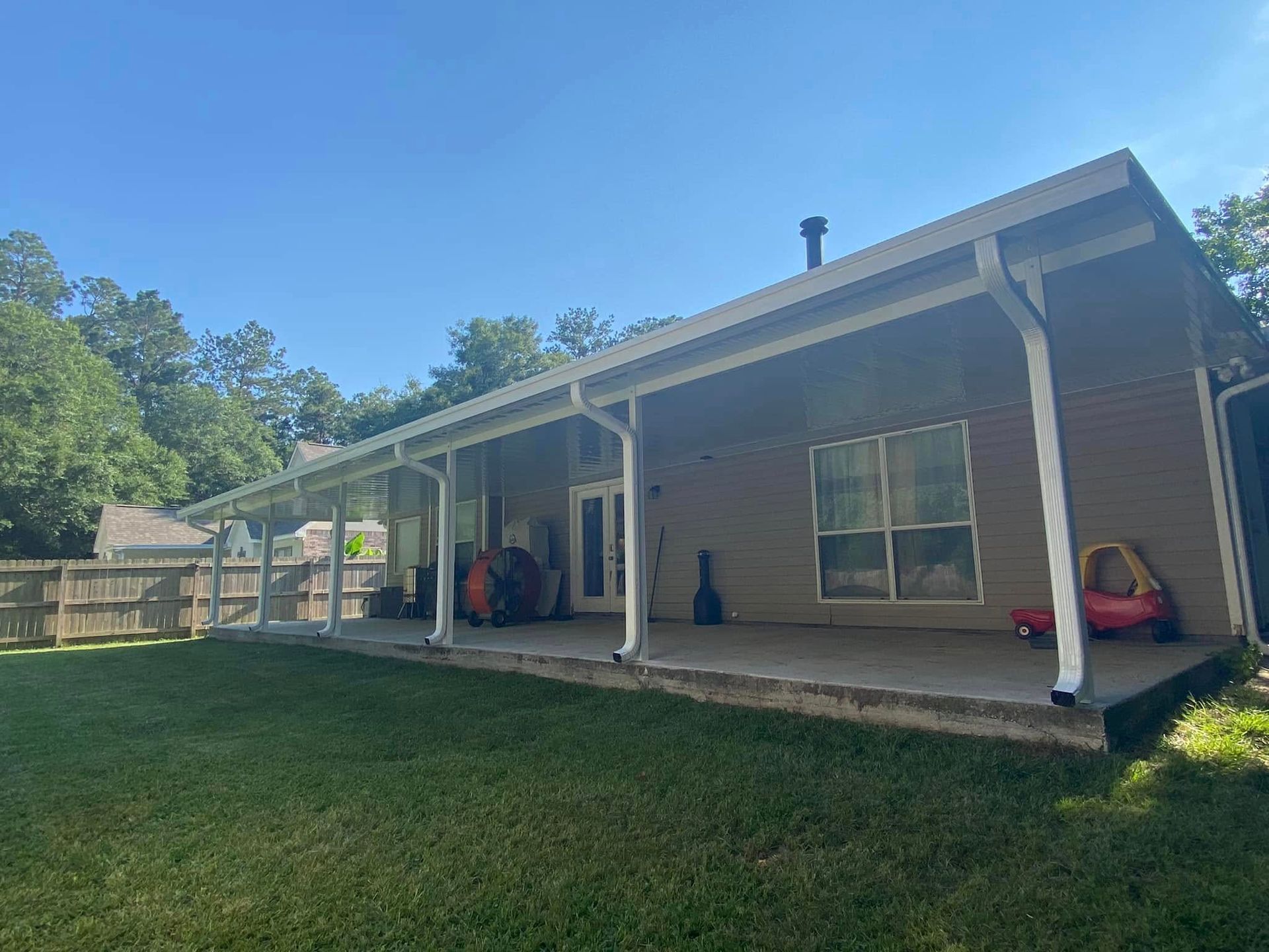 Backyard patio with roof, concrete floor, and brown siding. Green grass, blue sky, and a wooden fence.