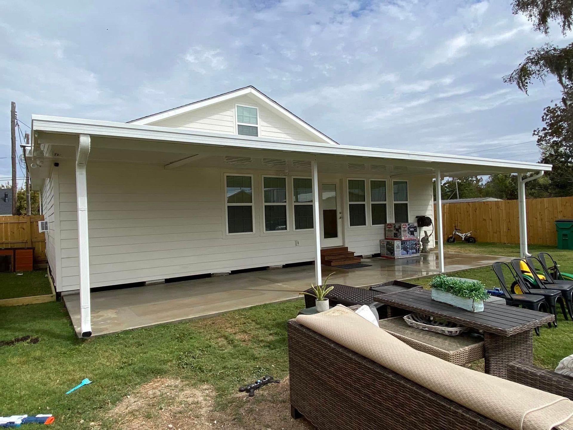 White house with a covered patio, featuring outdoor furniture on a green lawn under a cloudy sky.