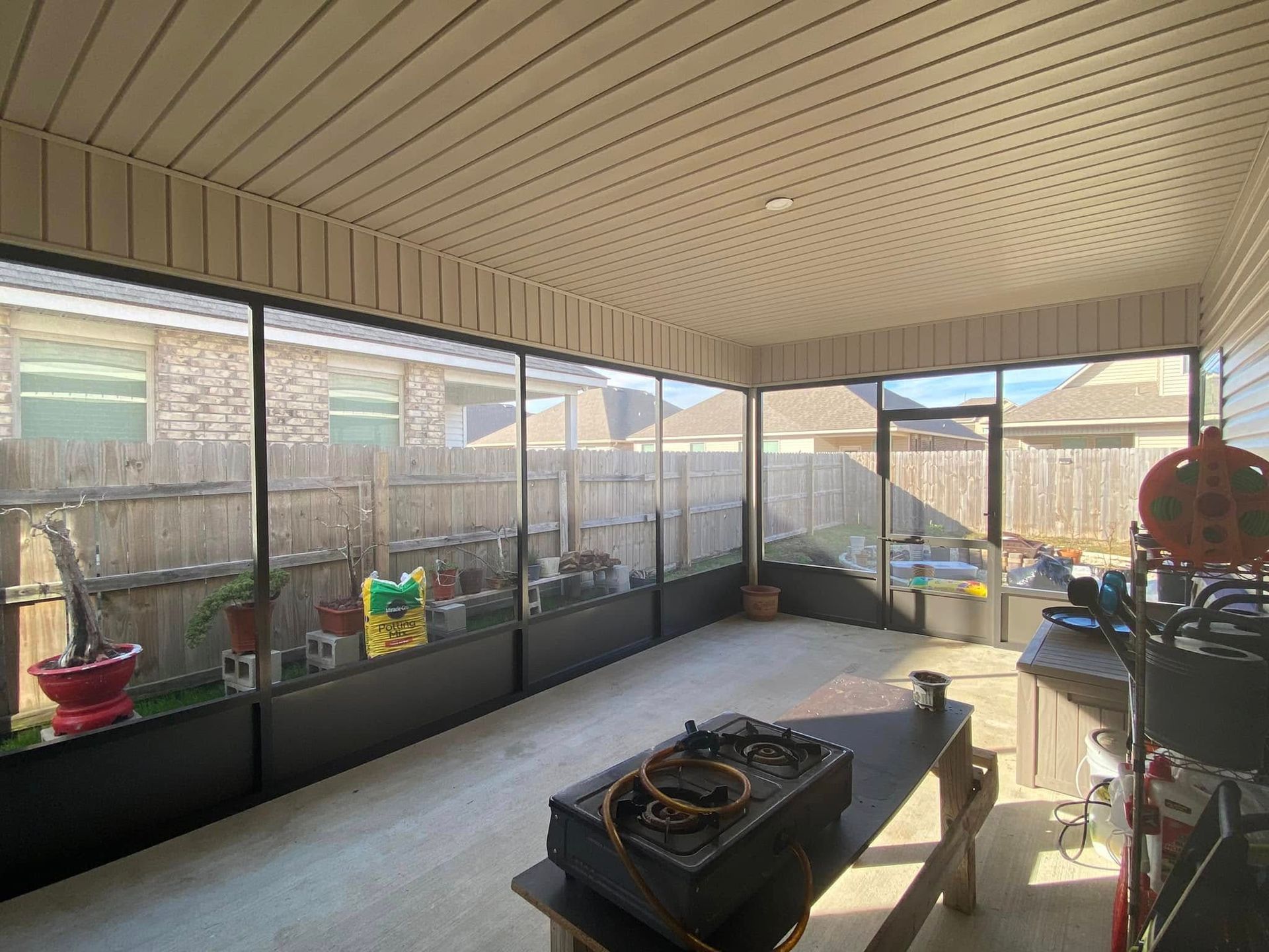 Screened-in porch with black frames, plants, a table, and a backyard view on a sunny day.