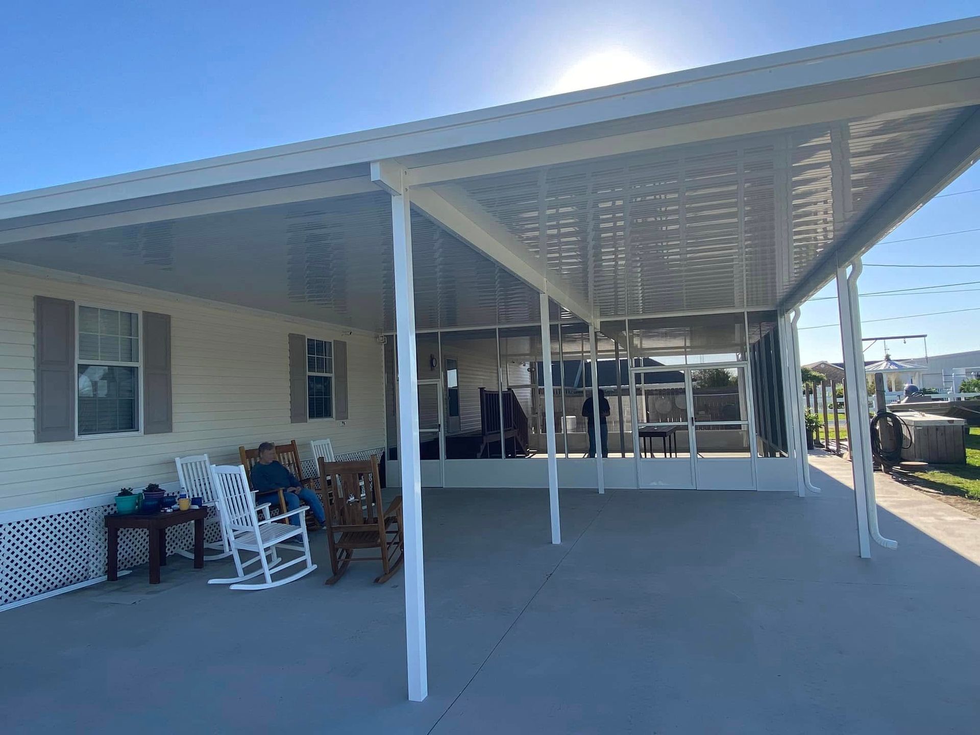 Covered porch with white support beams, attached to a cream-colored house. Rocking chairs sit on the gray patio.