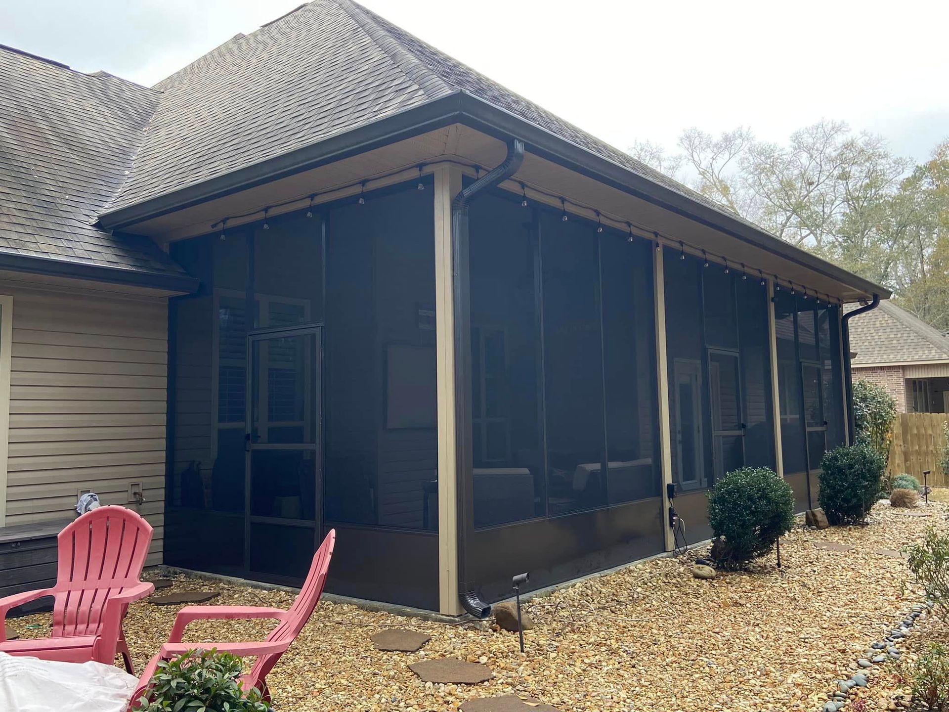 Screened-in porch attached to a house with black screens, tan trim, and gravel landscaping. Pink chairs are in the foreground.