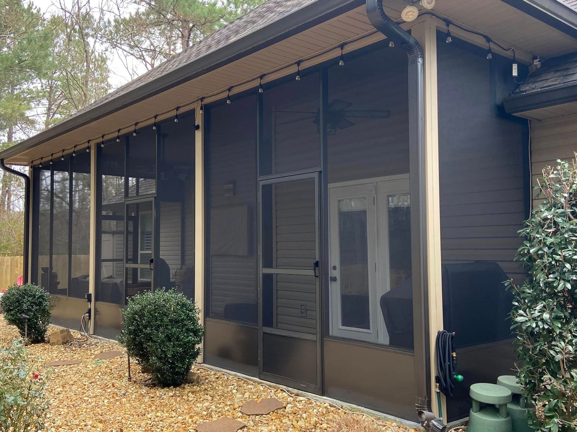 Screened-in porch with brown siding, a door, and bushes. Beige trim and string lights are visible.