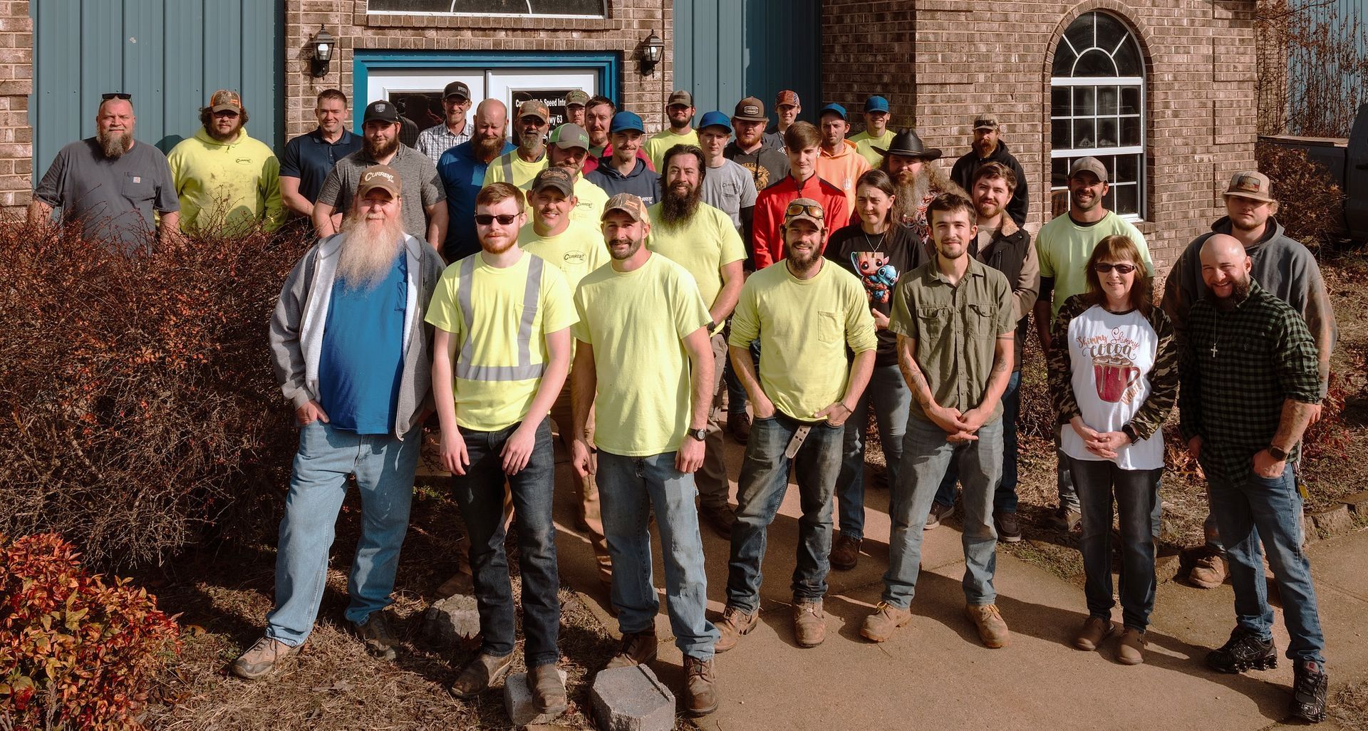 Group of construction workers in yellow shirts and jeans posing outside a brick building.
