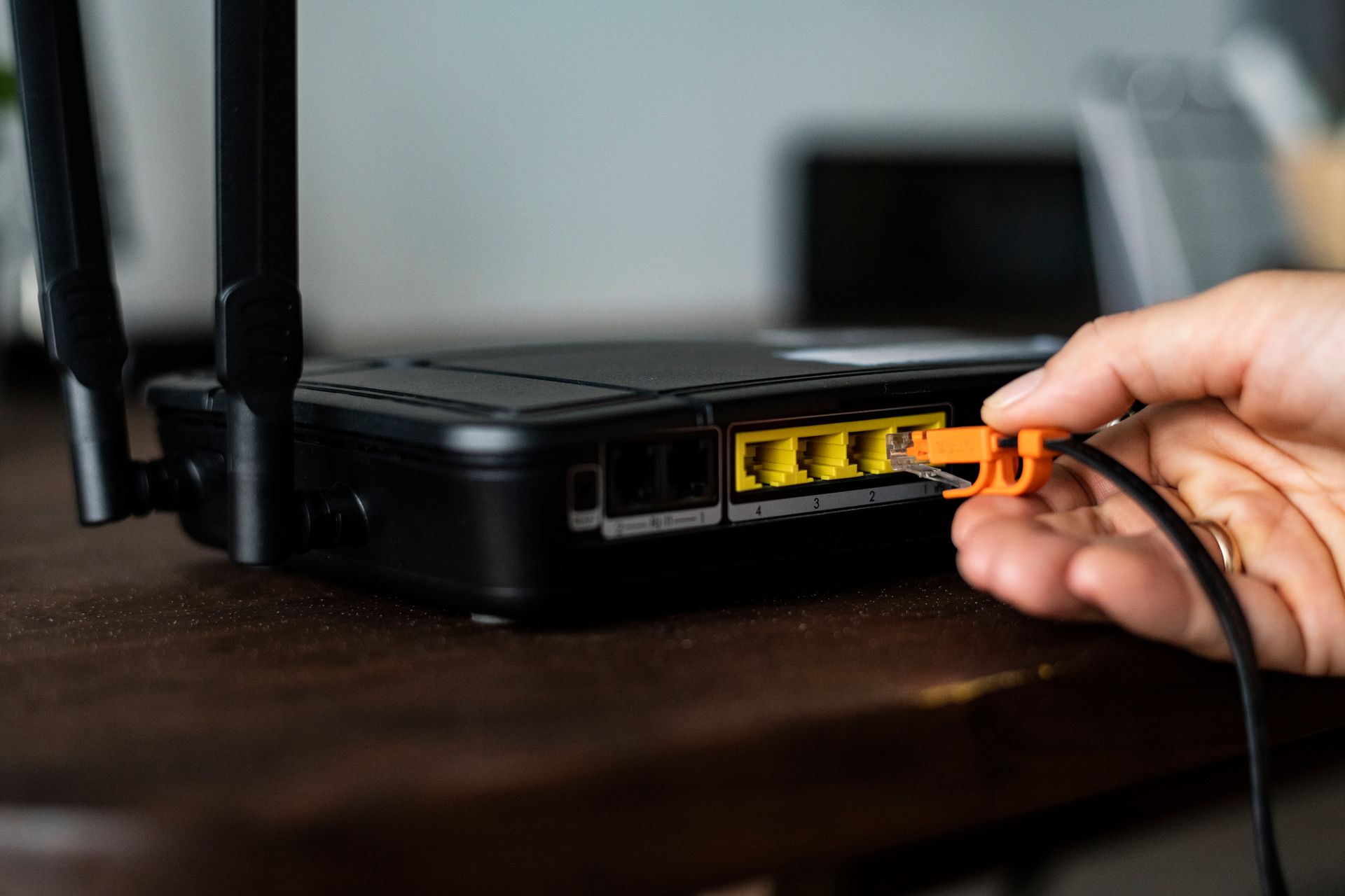 Hand plugging an orange Ethernet cable into a black router on a brown table.