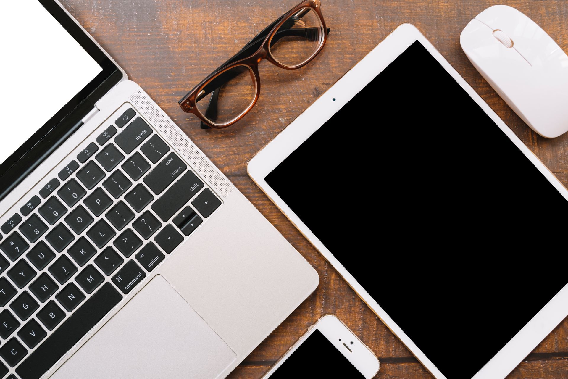 Laptop, tablet, phone, glasses, and mouse on a wood surface.