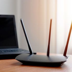 Black Wi-Fi router with three antennas next to a laptop on a wooden surface.