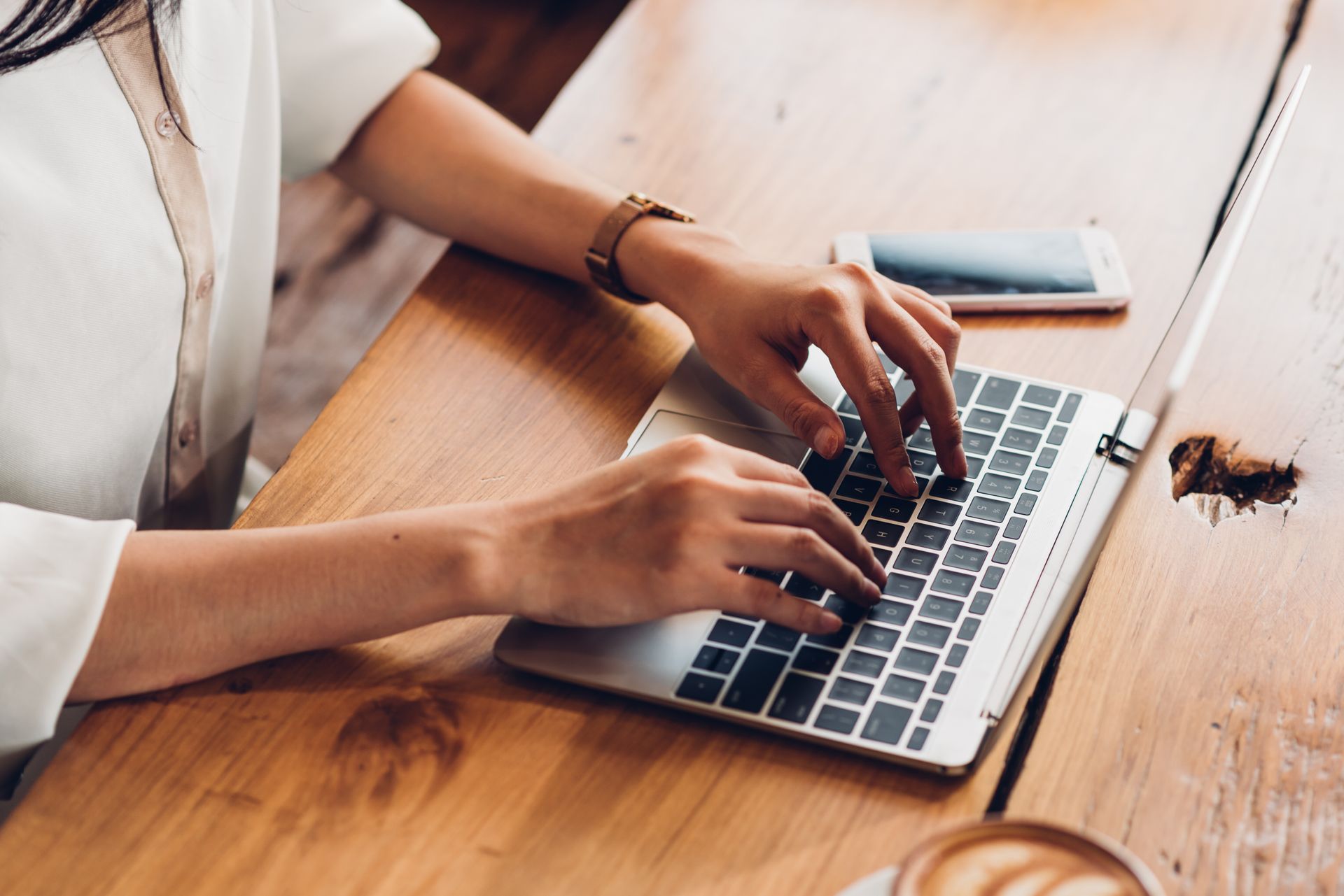 Woman's hands typing on a laptop at a wooden table, next to a smartphone and a coffee cup.