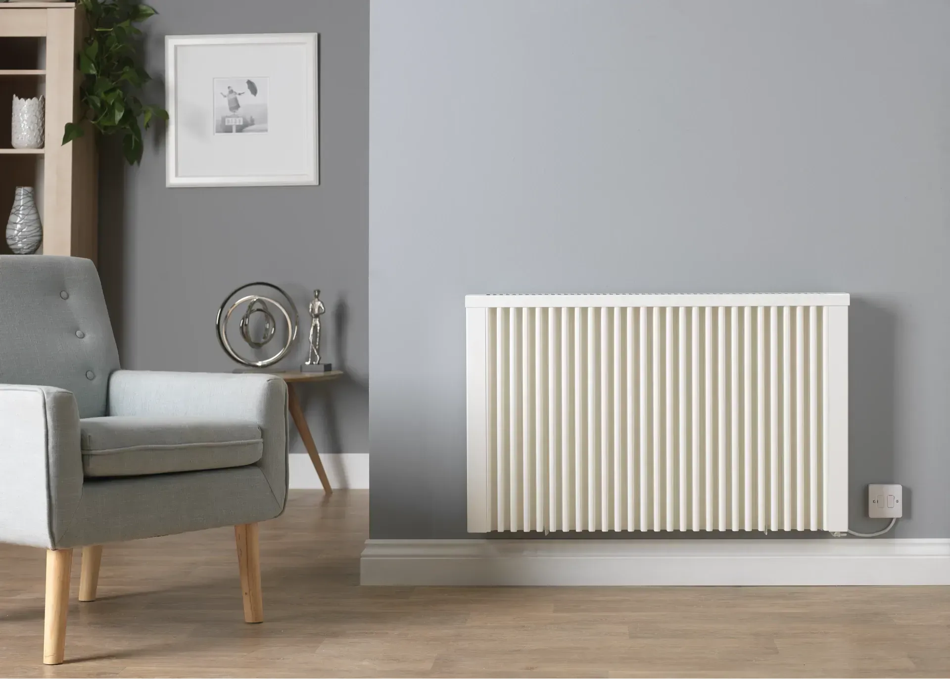 Wall-mounted cream radiator on a gray wall in a living room with an armchair, side table, and artwork.