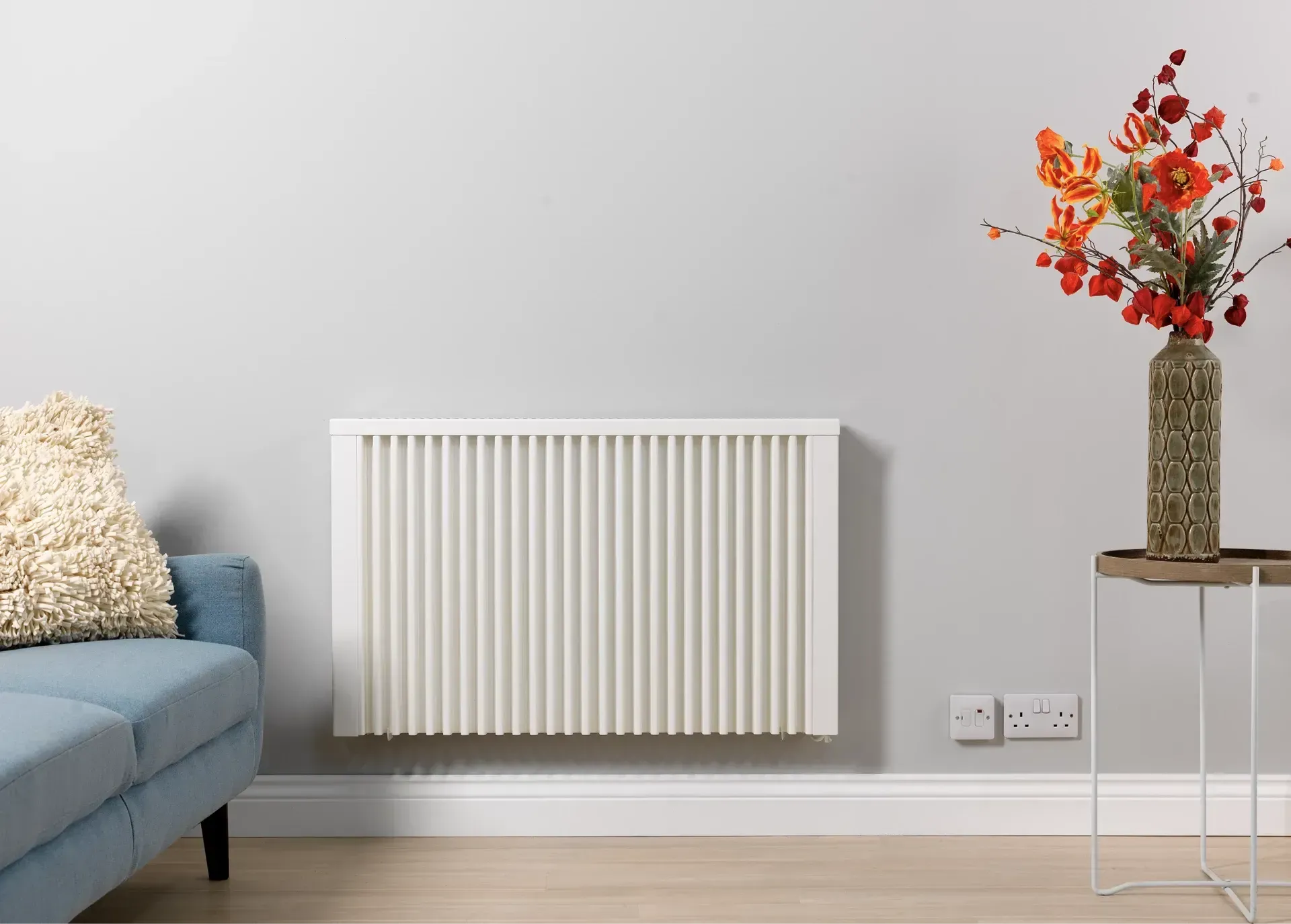 White wall-mounted radiator in a living room, next to a blue sofa and vase of flowers.