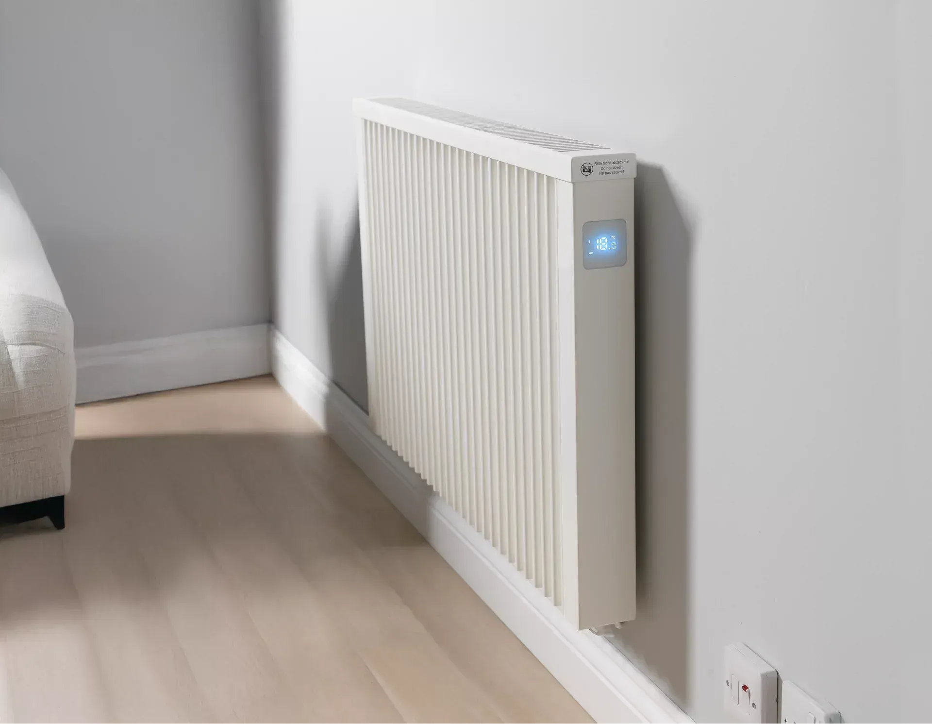 White wall-mounted electric radiator with blue light, next to a light-colored couch and power outlet.