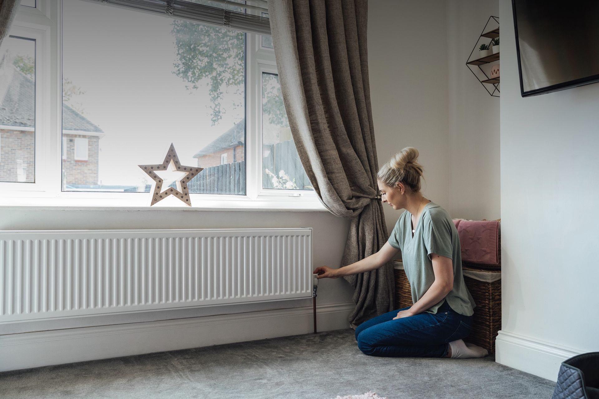 A woman is sitting on the floor in a living room looking at a radiator.