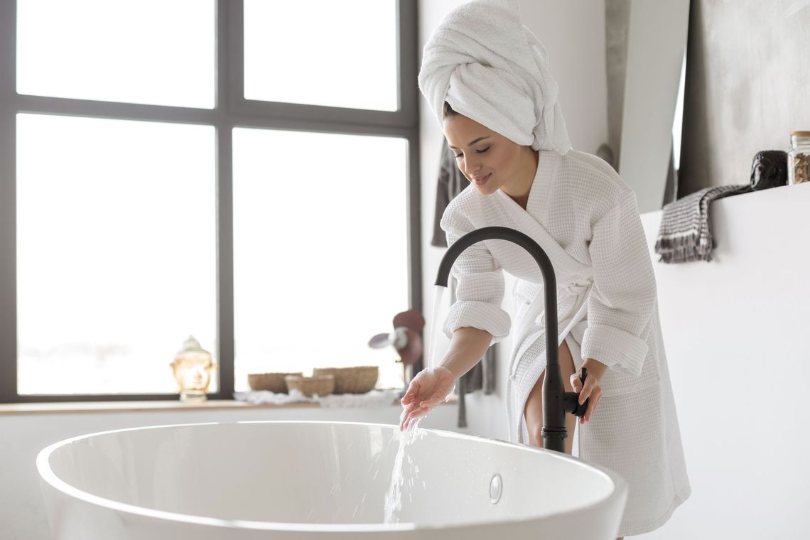 A woman in a bathrobe is pouring water into a bathtub.