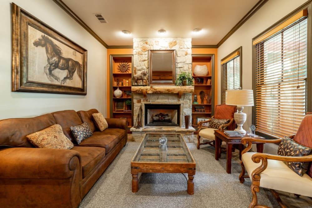 Cozy clubhouse interior with warm lighting featuring a large leather sofa, two armchairs with patterned cushions, a stone fireplace, and rustic decor. A large framed horse painting hangs on the wall, and the room is accented with wood furniture, decorative lamps, bookshelves, and horizontal blinds on the windows at Marquis at Bellaire Ranch in Fort Worth, TX.