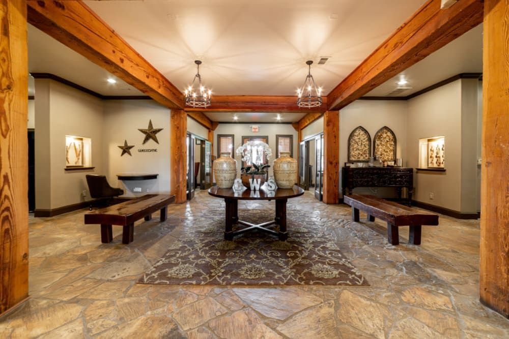 Elegant interior lobby area with rustic elements featuring exposed wooden beams and columns, ornate chandeliers, and a tile floor with a decorative pattern. Benches are positioned along the sides of the room, along with decorative wall elements including star sculptures and framed artwork. A reception desk can be seen in the distance with the word WELCOME displayed prominently at Marquis at Bellaire Ranch in Fort Worth, TX.