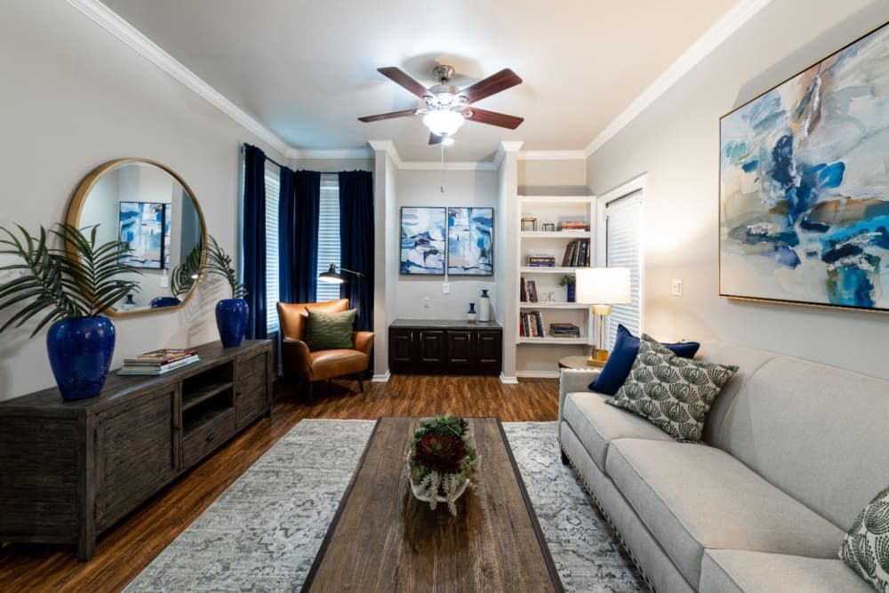 Cozy living room interior featuring modern furniture with a grey sofa and a brown leather armchair, accented by blue curtains and decor items. A stylish wooden entertainment unit sits opposite the sofa, flanked by vibrant blue vases and a circular mirror. The room is decorated with large abstract paintings with shades of blue, and a bookshelf filled with books adds a personal touch. A ceiling fan with wooden blades and a light adds warmth to the space, while plants bring a touch of greenery, completing the comfortable and inviting atmosphere at Marquis at Bellaire Ranch in Fort Worth, TX.