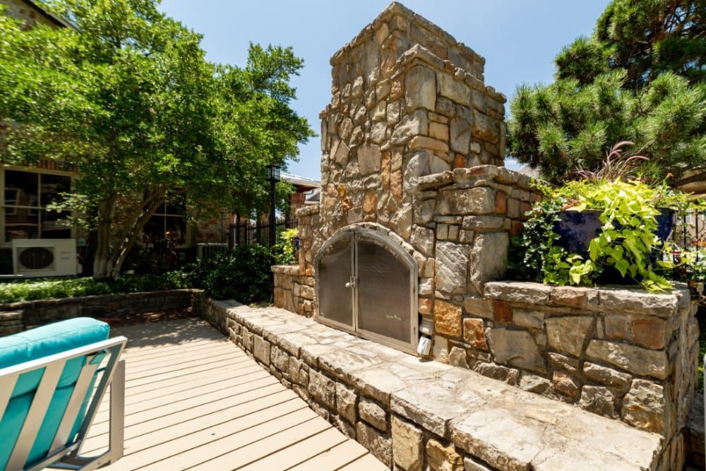 A sunny outdoor patio area featuring a tall stone fireplace with an arched opening and a metal screen. A wooden deck floor complements the warm hues of the stonework. Lush greenery, including a well-lit tree and potted plants, adds a tranquil vibe to the space. Part of a turquoise-striped lounge chair is visible in the foreground, implying a comfortable place to relax at Marquis at Bellaire Ranch in Fort Worth, TX.
