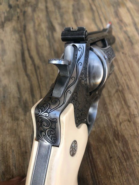Close-up of a revolver with intricate floral engravings on the steel frame and ivory-colored grip against a wood background.