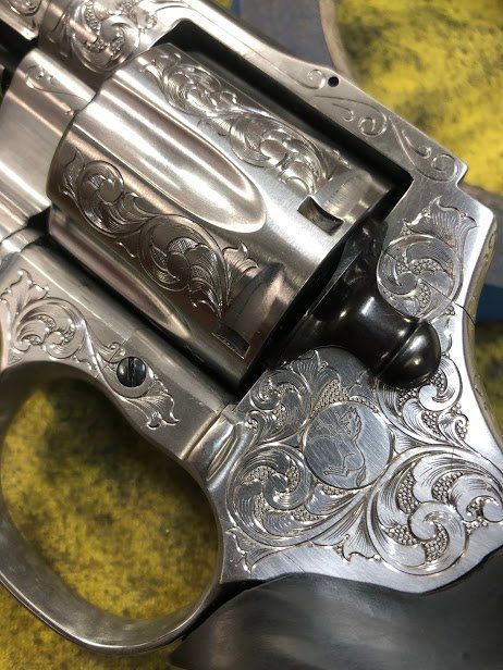 Close-up of engraved silver revolver with intricate floral patterns. Cylinder open, revealing chambers.