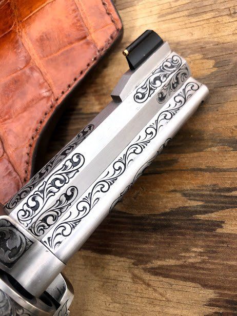 Close-up of an engraved silver handgun. Decorative scrollwork along the barrel, set against a leather holster.