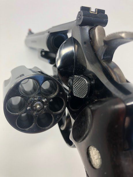 Close-up of a black revolver with the cylinder open, showing six chambers. Dark wood grip.