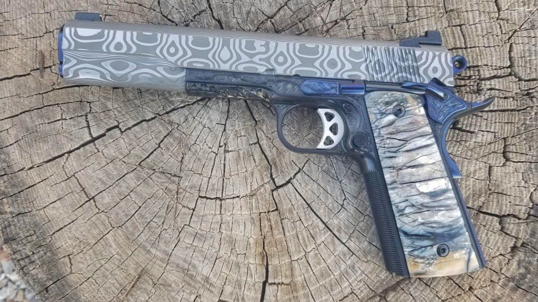 Custom 1911 pistol with Damascus steel slide and camo grip, resting on a wooden surface.