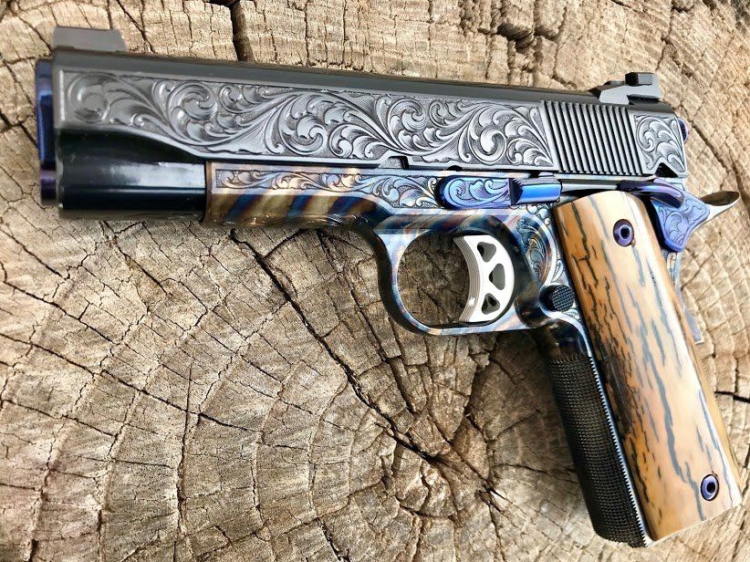 Custom engraved 1911 pistol with blue accents and wood-grain grips, resting on a wood stump.