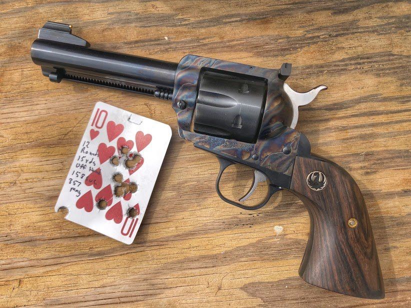 Revolver with a patterned finish next to a playing card riddled with bullet holes on a wooden surface.