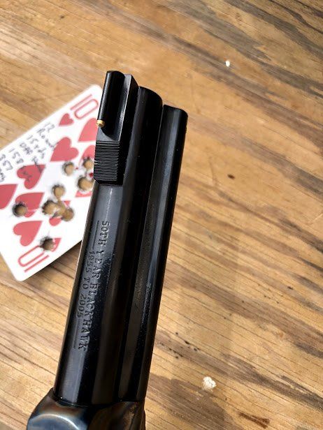 A dark handgun barrel, sights aligned, with a playing card target marked with bullet holes in the background.