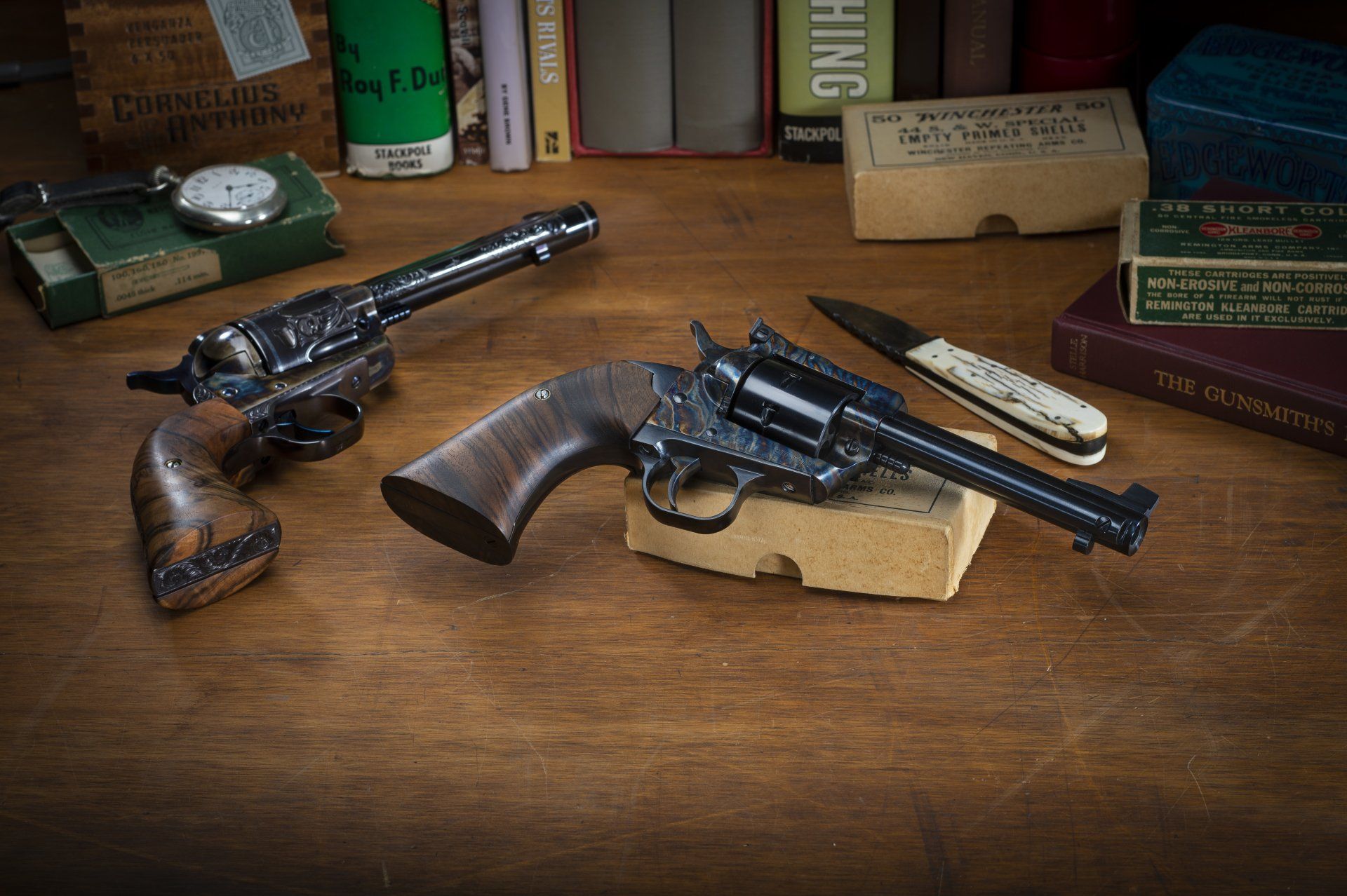 Two antique revolvers on a wooden table with a pocket knife, books, and vintage boxes.
