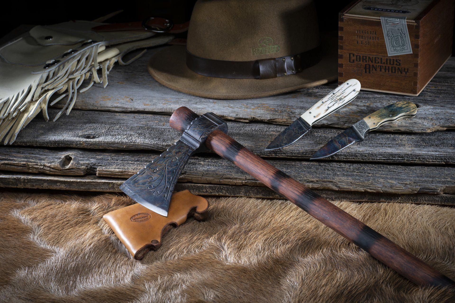 Axe, hat, knives, and box on a rustic surface. Browns and tans dominate, suggesting an outdoors or hunting theme.
