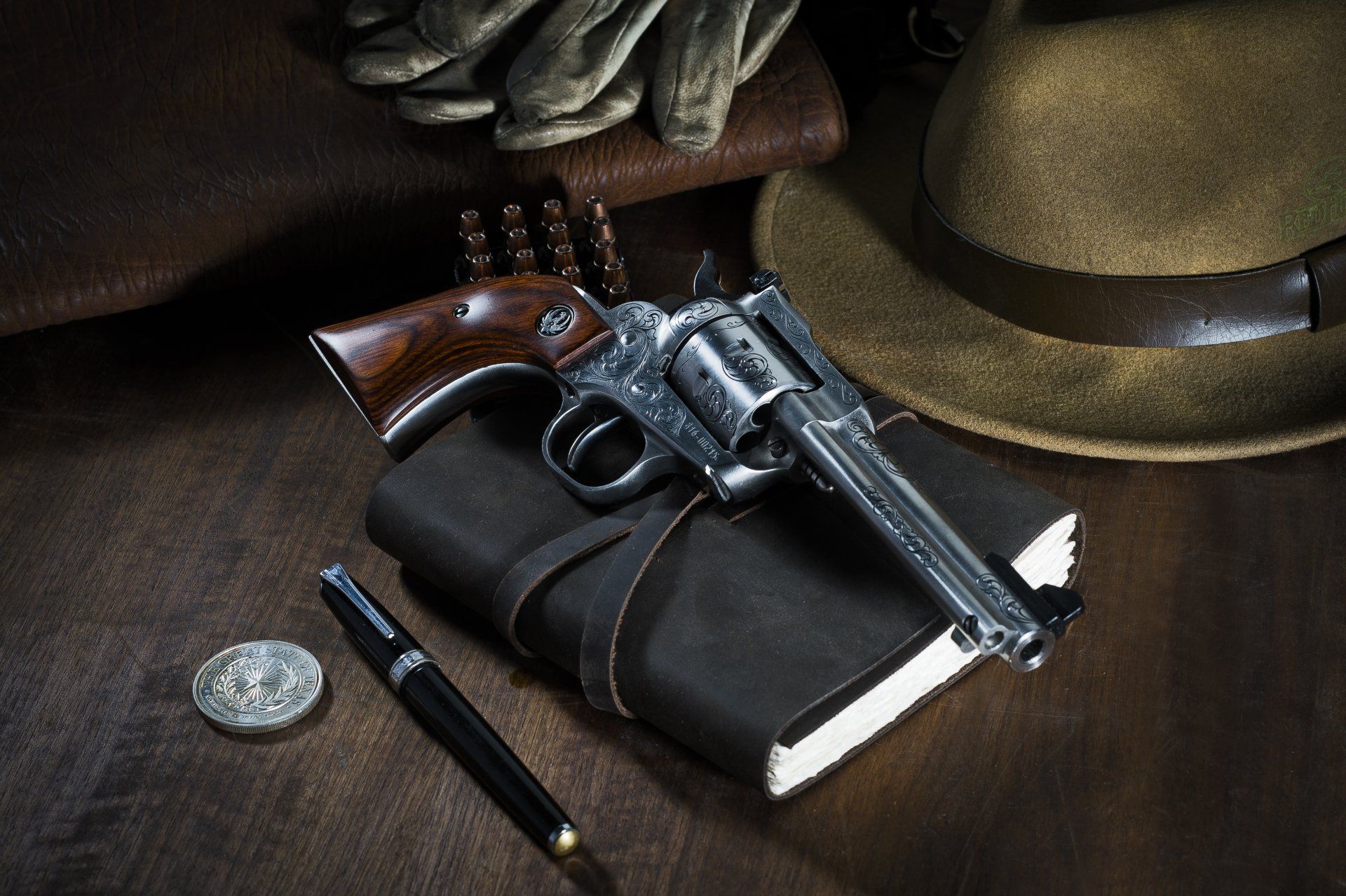 Revolver gun on a leather-bound book, with a hat, gloves, coin, and pen on a wooden surface.