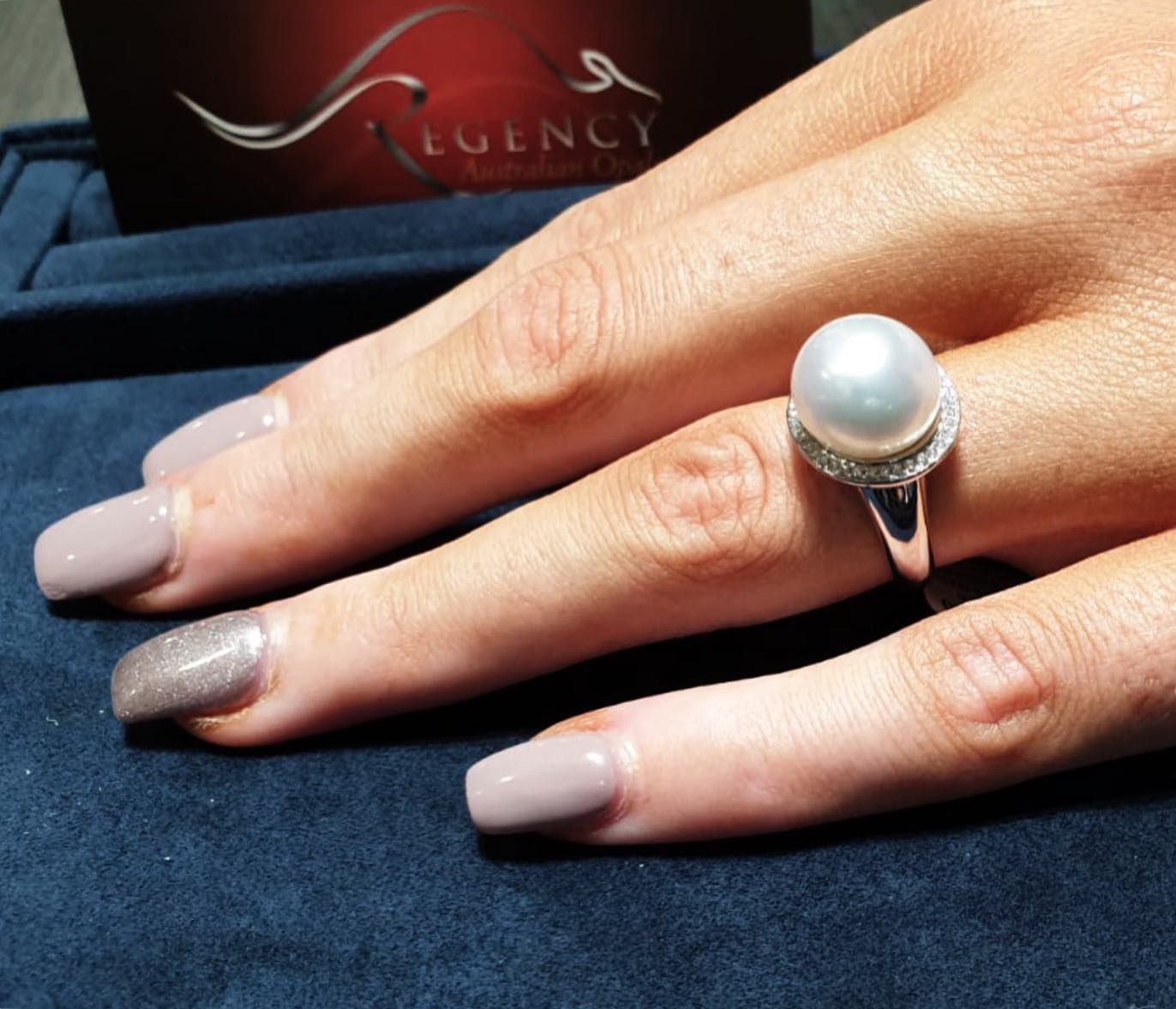 Hand Wearing a Pearl Ring With a Diamond Halo on a Blue Surface — Regency Jewellers in Cairns City, QLD