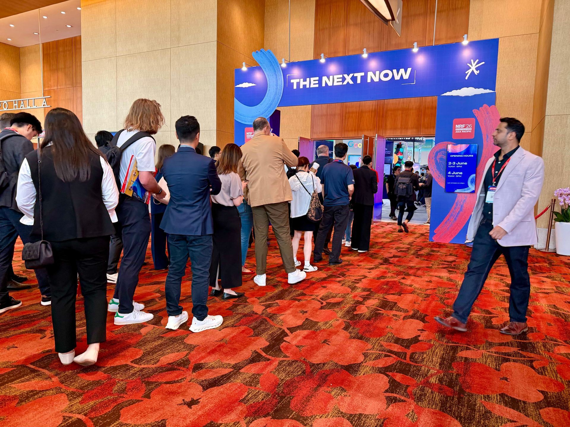 People waiting in line at a conference entrance with a blue “THE NEXT NOW” sign.