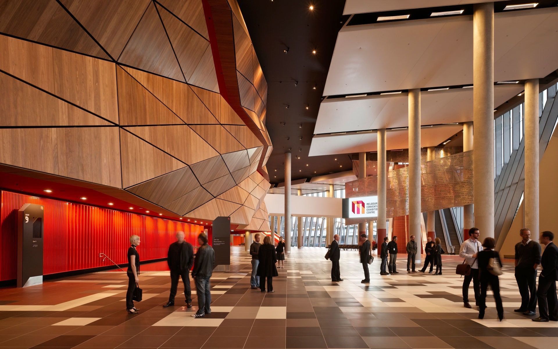 Interior of a modern building lobby with people, geometric wood and metal walls, pillars, and red curtains.