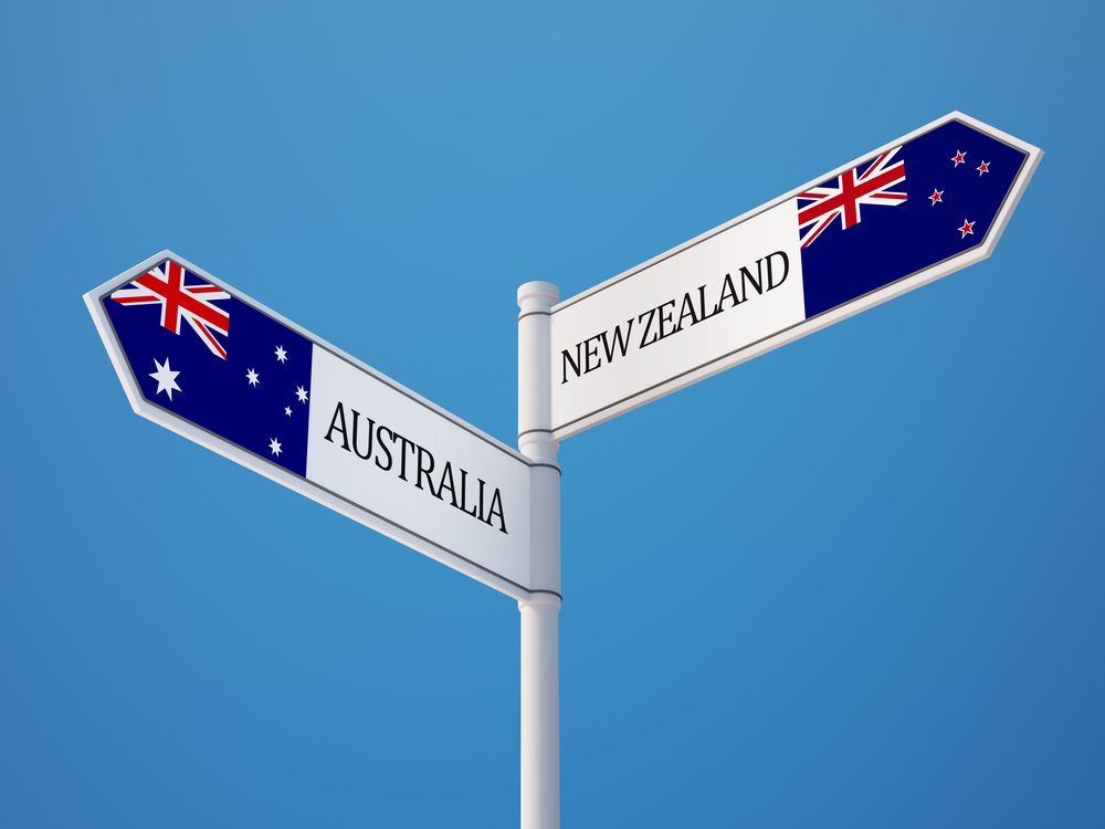 Signpost with flags for Australia and New Zealand, pointing in opposite directions against a blue sky.