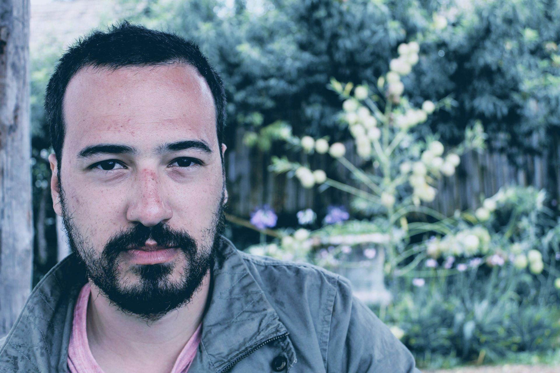 A man with a beard is looking at the camera with flowers in the background