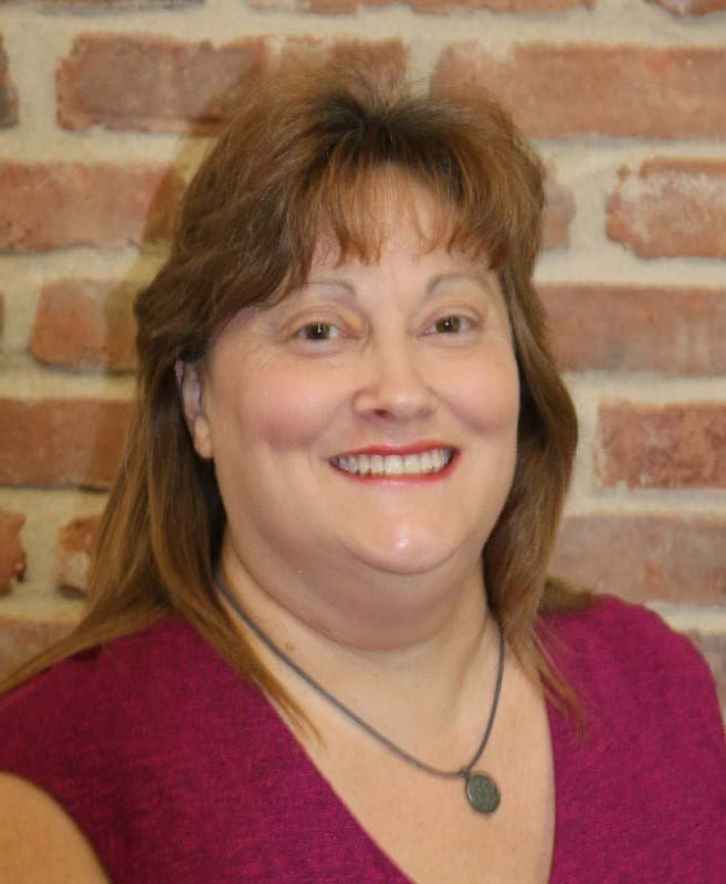 A woman in a purple shirt is smiling in front of a brick wall