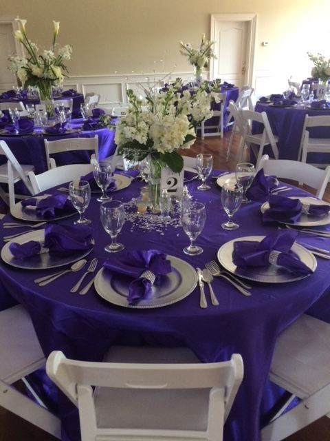A table set for a wedding reception with purple table cloths and white chairs