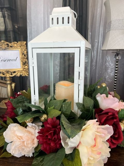 A white lantern is surrounded by flowers and leaves on a table.