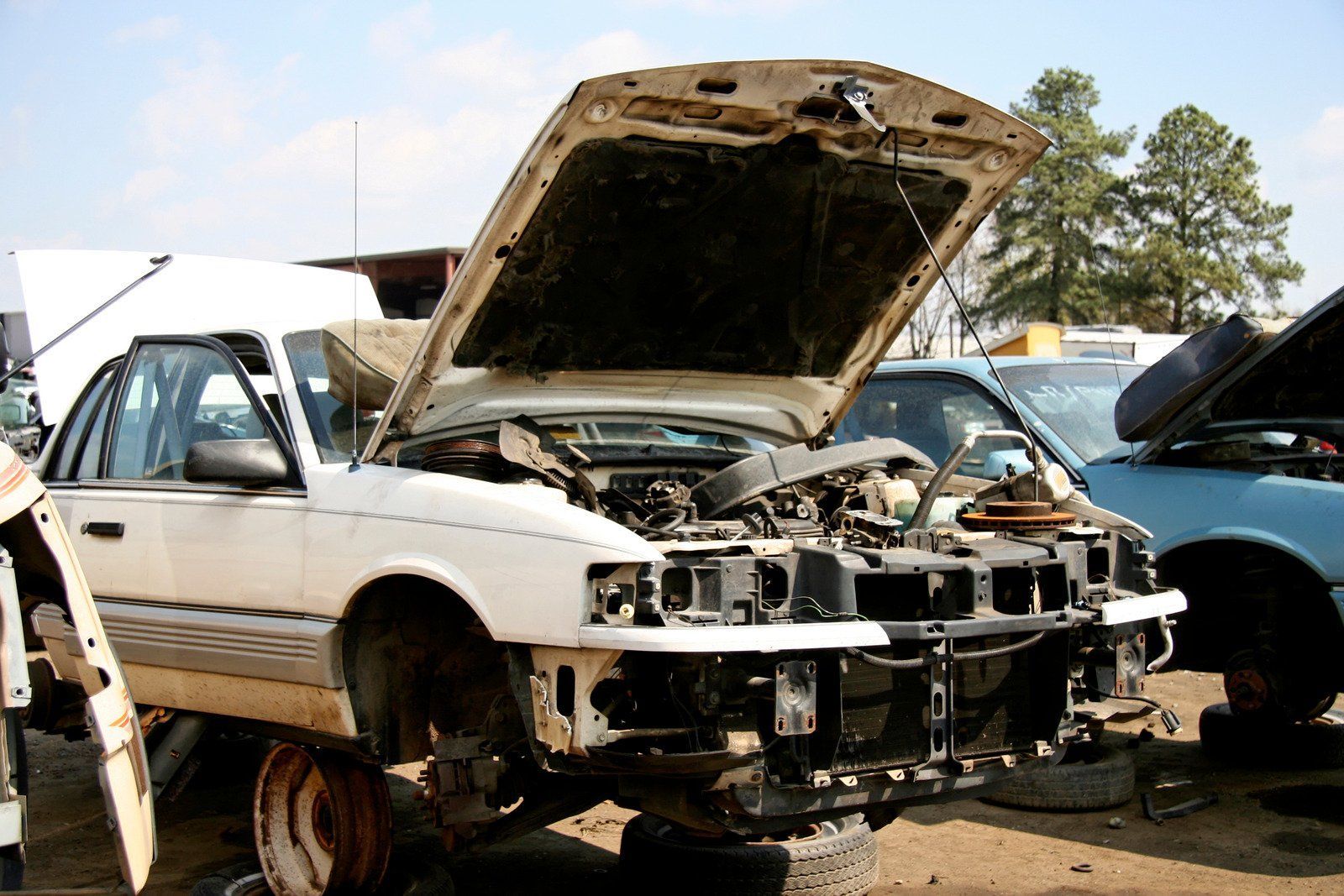 White car in a junkyard with hood open, front end missing, and other damaged cars in the background.
