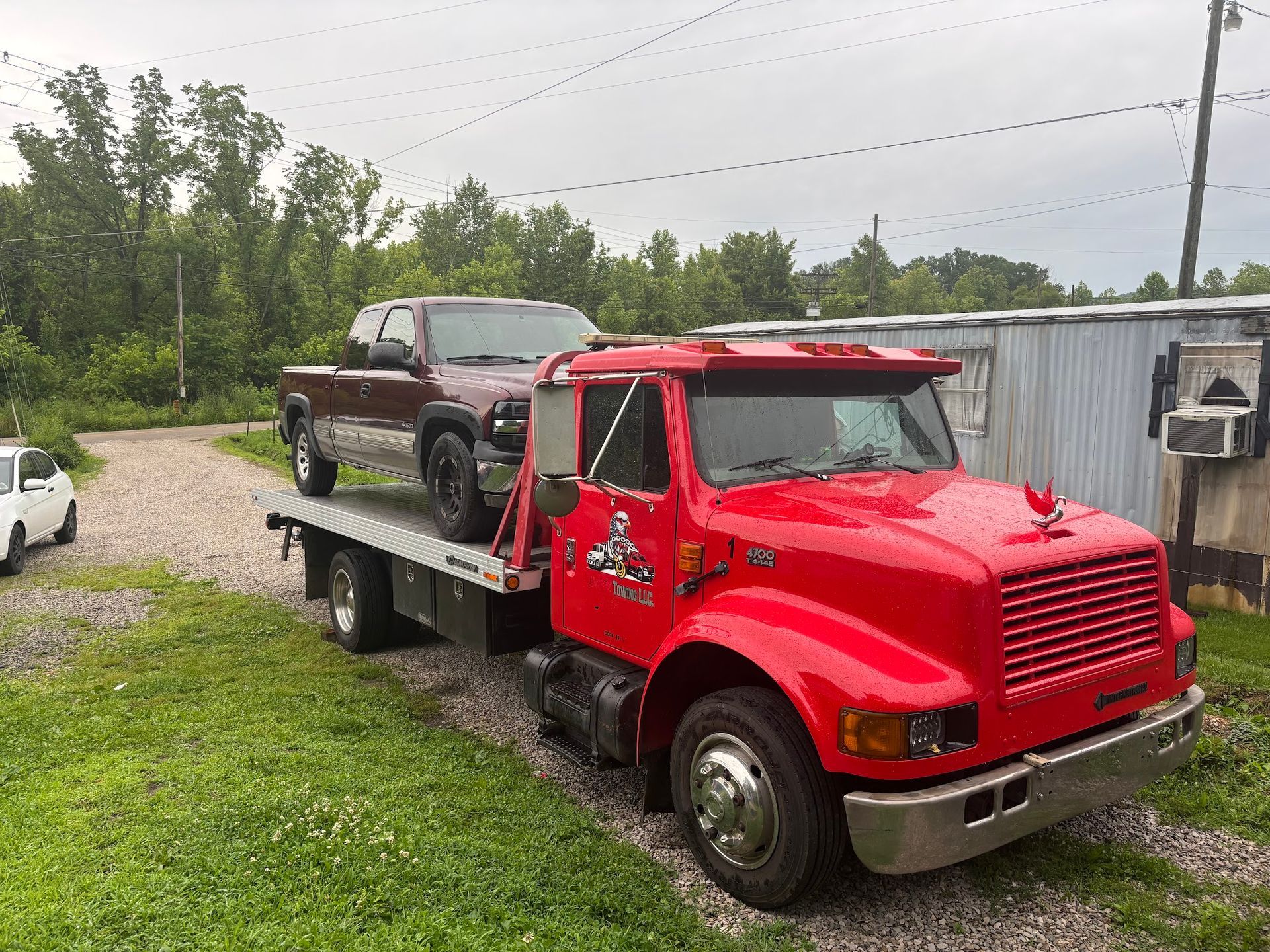 Red tow truck hauling a brown pickup truck on a grassy driveway near a building.