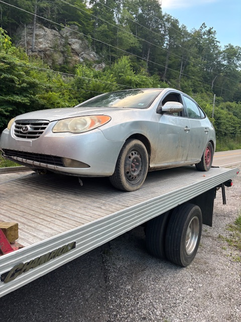 Silver Hyundai Elantra on a tow truck, parked on a dirt road, with a forested hill in the background.